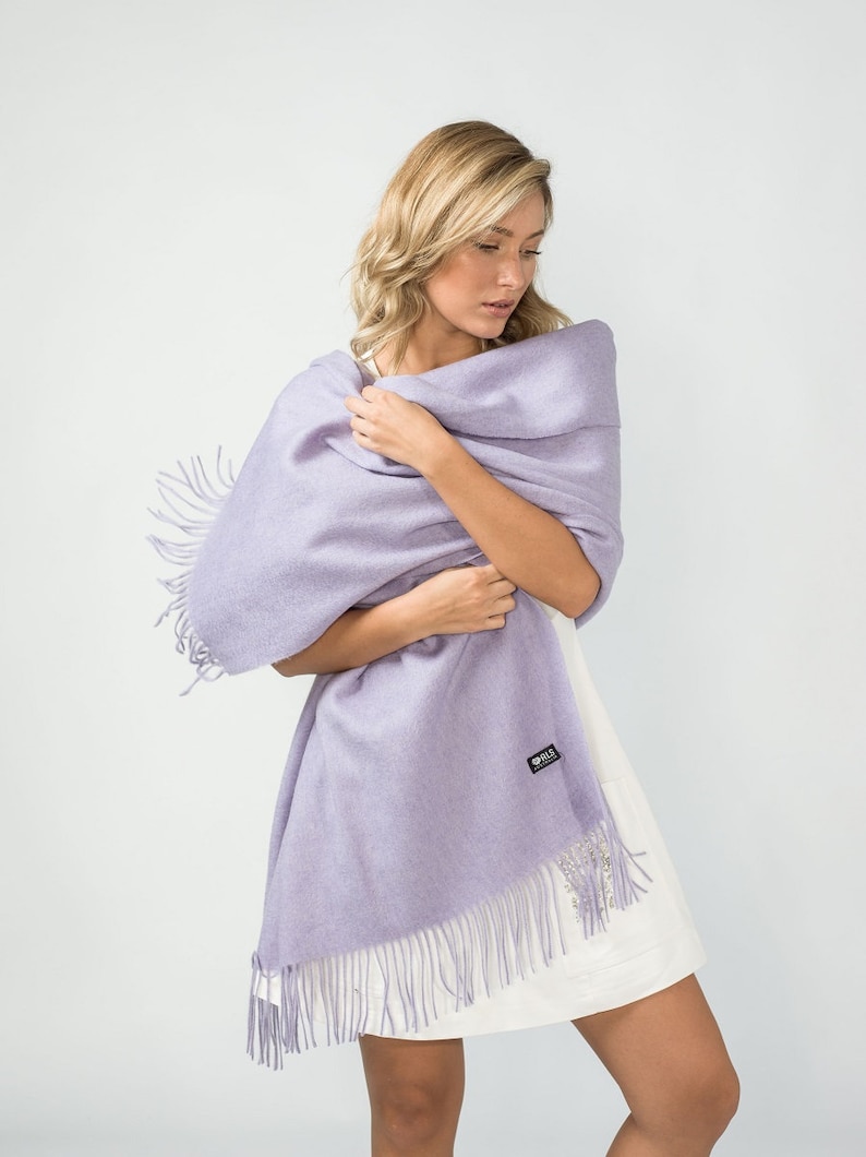 May include: A lavender pashmina shawl with fringe. The shawl is draped over a person's shoulders and is held in place by their hands.