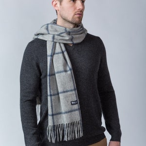 May include: A grey and blue plaid scarf with fringe. The scarf is worn around the neck of a person wearing a dark grey sweater and brown trousers.