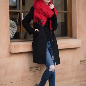 May include: A woman wearing a black coat, red scarf, blue jeans, and green suede boots with gold accents. She is standing next to a window.
