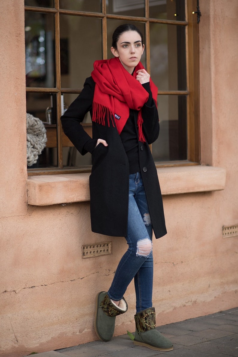 May include: A woman wearing a black coat, red scarf, blue jeans, and green suede boots with gold accents. She is standing next to a window.