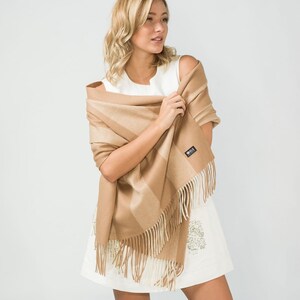 May include: A woman wearing a white dress is holding a brown and beige striped scarf with fringe. The scarf has a small black label with the word "Silk" on it.