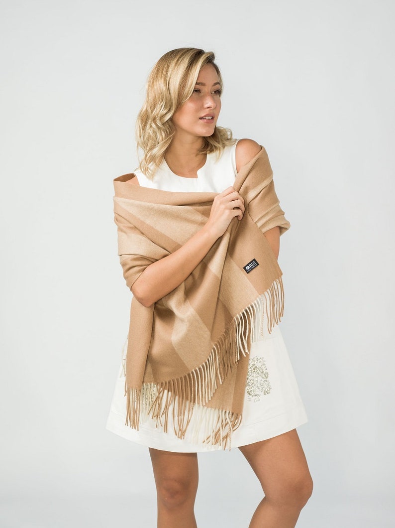 May include: A woman wearing a white dress is holding a brown and beige striped scarf with fringe. The scarf has a small black label with the word "Silk" on it.