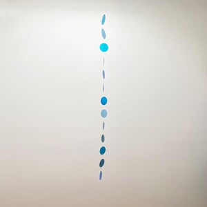 May include: A hanging mobile featuring a vertical string of blue, circular shapes in varying shades. The shapes range from light to dark blue, creating a gradient effect. The mobile is suspended against a plain white background.