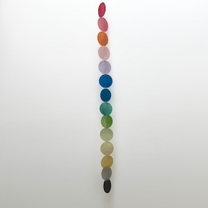 May include: A vertical hanging garland consisting of a single column of circular discs arranged in a gradient of colours. The discs transition from shades of pink and orange at the top, through blue and green in the middle, to yellow, grey, and black at the bottom.