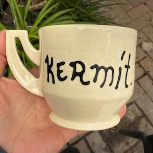Puede incluir: Taza de cerámica color crema con la palabra "Kermit" en letras negras. La taza tiene un diseño de asa único y una superficie ligeramente texturizada. Un artículo de cocina y comedor.