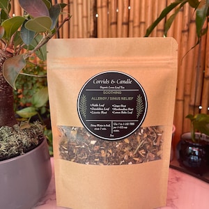 Puede incluir: Una bolsa de papel marrón con una ventana transparente que muestra una mezcla de hierbas secas. La bolsa está etiquetada "Corvids & Candle Organic Loose Leaf Tea SOOTHING ALLERGY/SINUS RELIEF" y enumera los ingredientes: hoja de ortiga, hoja de diente de león, raíz de regaliz, raíz de jengibre, raíz de malvavisco y hoja de melisa. La bolsa también incluye instrucciones para preparar el té: "Llevar el agua a ebullición, dejar reposar 7 min. Usar 1 a 1 1/2 cucharadas por 1 1/2 taza de agua. 2oz"