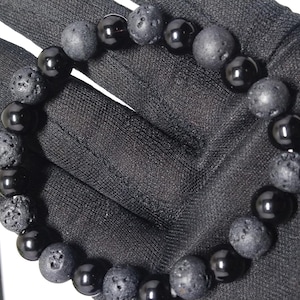 May include: A black beaded bracelet featuring a combination of smooth, polished onyx beads and textured lava stone beads. The bracelet is displayed on a black surface, showcasing the contrast between the materials.