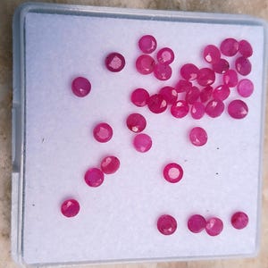 May include: A collection of small, round, faceted loose rubies with a vibrant pinkish-red hue. These gemstones are arranged on a white foam insert inside a square, clear plastic display box, showcasing their reflective surfaces and uniform shape.
