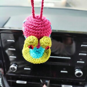 Pu&ograve; includere: Un ciondolo per auto fatto a mano, all'uncinetto a forma di creatura stravagante. Il ciondolo presenta un corpo rosa, dettagli gialli e blu e dettagli rossi. &Egrave; appeso a un cordino rosa, aggiungendo un tocco di colore.