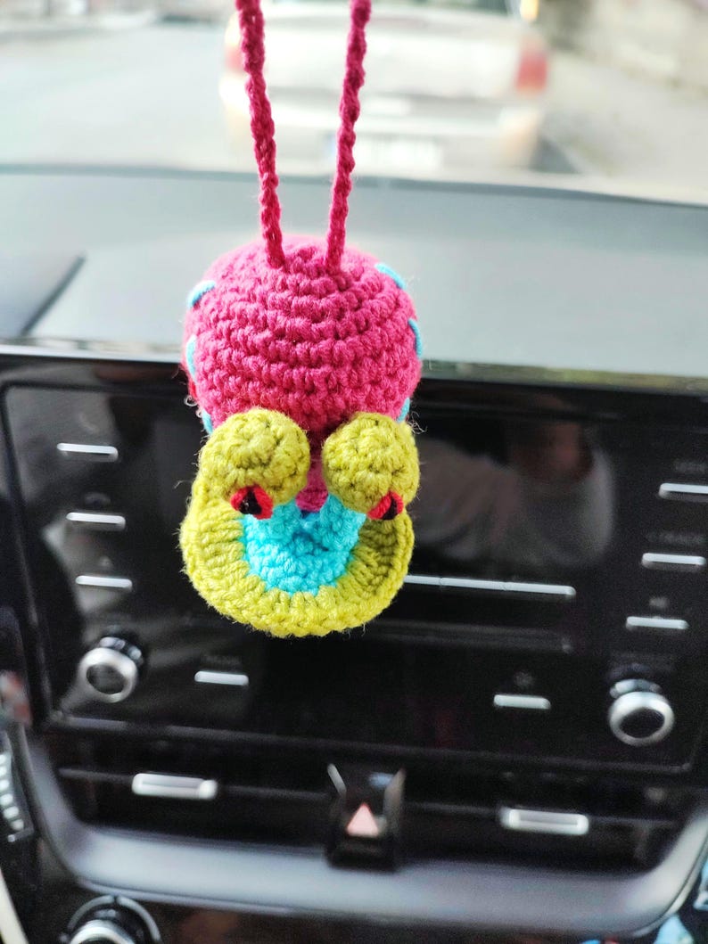 Pu&ograve; includere: Un ciondolo per auto fatto a mano, all'uncinetto a forma di creatura stravagante. Il ciondolo presenta un corpo rosa, dettagli gialli e blu e dettagli rossi. &Egrave; appeso a un cordino rosa, aggiungendo un tocco di colore.