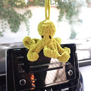 Op de afbeelding: Een limoengroene gehaakte octopus die aan een achteruitkijkspiegel van een auto hangt. De pluche octopus heeft een textuur van garen, een ronde kop en acht gekrulde tentakels. Hij hangt aan een bijpassende groene lus van garen aan de spiegel.