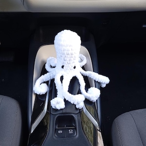 Op de afbeelding: Een wit gehaakt octopus speelgoed met acht tentakels. Het pluche speelgoed staat op de middenconsole van een auto. De octopus heeft twee zwarte ogen en een ronde kop. De tentakels zijn gekruld en zacht.