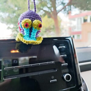 Pu&ograve; includere: Un ciondolo per auto fatto a mano all'uncinetto a forma di lumaca dei cartoni animati. La lumaca ha un corpo viola, grandi occhi gialli con dettagli rossi e un guscio blu e verde. &Egrave; appeso a un cordino viola.