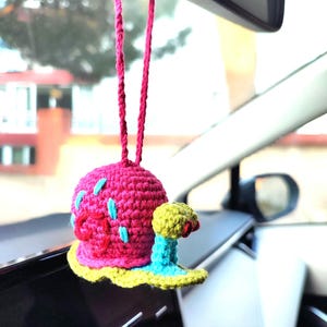 Pu&ograve; includere: Un ciondolo per auto fatto a mano all'uncinetto raffigurante una lumaca rosa con dettagli blu e un fiore giallo e rosa. Il ciondolo &egrave; appeso a un cordino rosa, aggiungendo un tocco di fantasia all'interno di un veicolo.