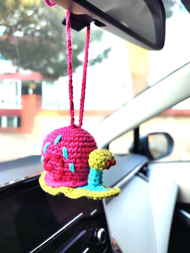 Pu&ograve; includere: Un ciondolo per auto fatto a mano all'uncinetto raffigurante una lumaca rosa con dettagli blu e un fiore giallo e rosa. Il ciondolo &egrave; appeso a un cordino rosa, aggiungendo un tocco di fantasia all'interno di un veicolo.