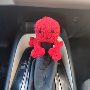 Può includere: Un giocattolo a forma di polpo rosso brillante, lavorato all'uncinetto, con un volto sorridente e occhi neri. Il polpo è attaccato alla leva del cambio di un'auto. Il peluche è un accento vivace all'interno del veicolo.