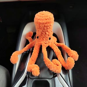 Könnte beinhalten: Ein leuchtend orangefarbenes, gehäkeltes Oktopus-Stofftier mit strukturierter Garnoberfläche, bestehend aus einem abgerundeten Kopf und acht langen, geringelten Tentakeln. Der Oktopus hat zwei kleine, dunkle, runde Augen auf der Vorderseite.
