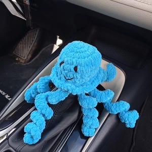 Peut inclure: Une peluche pieuvre crochetée à la main, de couleur bleu clair, avec un visage souriant et des yeux marron foncé. La pieuvre a huit tentacules bouclés et repose sur une surface noire. Fabriqué en fil doux et texturé.