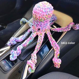 Op de afbeelding: Een veelkleurige gehaakte octopus, voornamelijk roze, paars en blauw. Het speelgoed heeft acht lange, getextureerde tentakels en is gelabeld met "3411 color". De octopus ligt op de console van een auto.