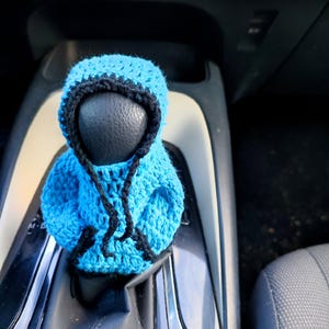 May include: A blue crocheted hooded figure with black trim, used as a gear shift knob cover in a car.