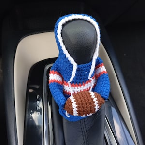 Op de afbeelding: Een blauw, wit en rood gehaakte hoodie met een bruine en witte voetbal. De hoodie is ontworpen om over de versnellingspookknop van een auto te passen. De voetbal wordt in de armen van de hoodie gehouden.