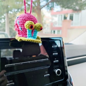 Pu&ograve; includere: Un ciondolo per auto a forma di lumaca all'uncinetto. La lumaca ha un guscio rosa con dettagli blu e rossi, occhi verdi e gialli e steli oculari blu. &Egrave; appeso a un filo rosa davanti a un cruscotto nero.