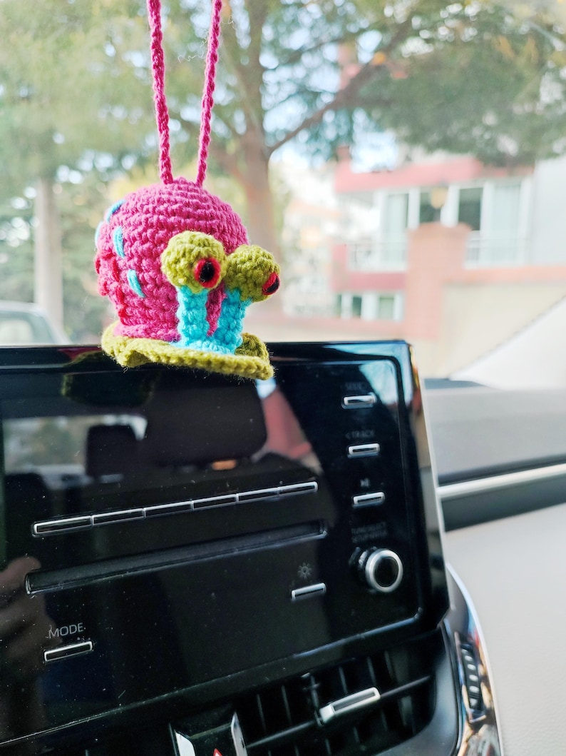 Pu&ograve; includere: Un ciondolo per auto a forma di lumaca all'uncinetto. La lumaca ha un guscio rosa con dettagli blu e rossi, occhi verdi e gialli e steli oculari blu. &Egrave; appeso a un filo rosa davanti a un cruscotto nero.