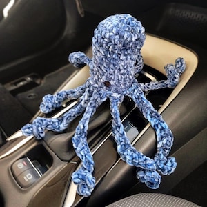 May include: A blue and white crocheted octopus toy with eight tentacles. The plush toy is made of a soft, textured yarn and is sitting on a car console.