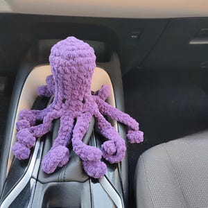 May include: A handmade, plush purple octopus toy. The octopus has a rounded head and eight long, curled tentacles. The toy is made of a soft, textured material and is resting on a car's gear shift.