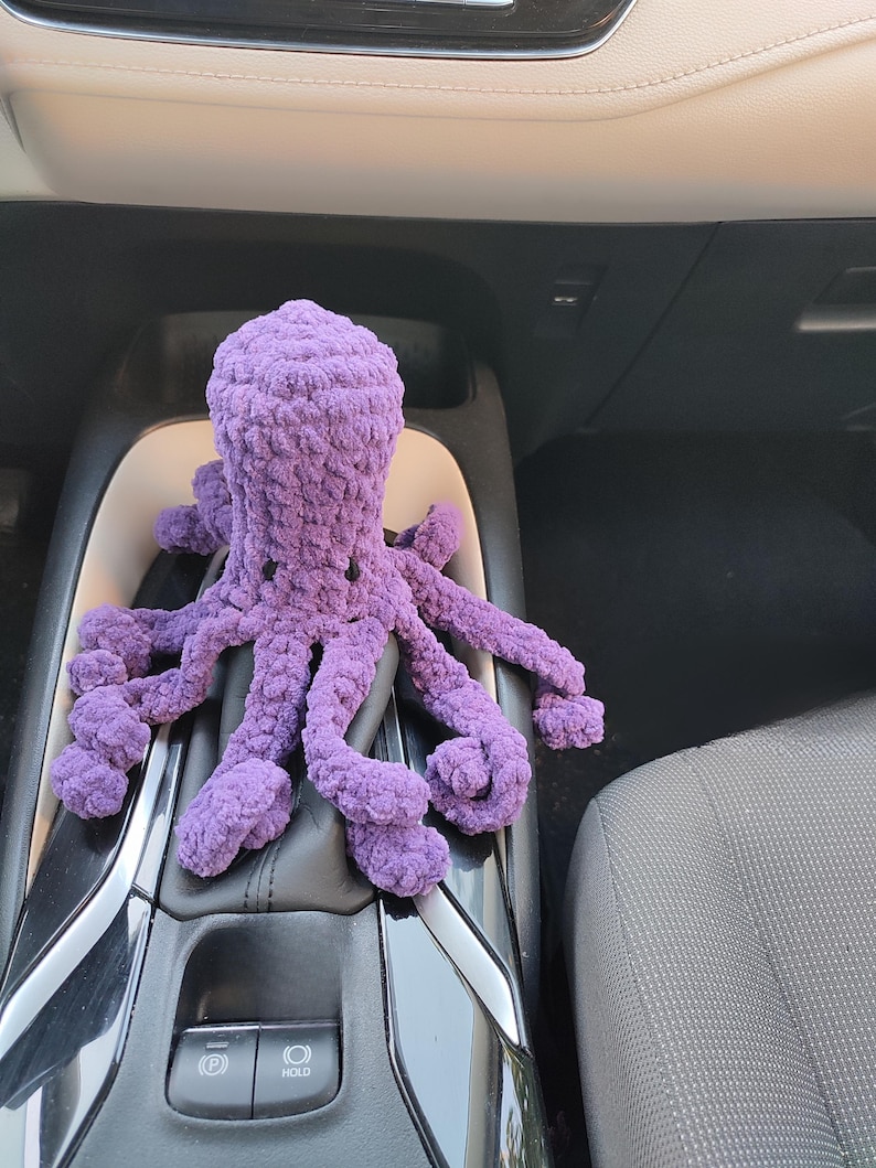 May include: A handmade, plush purple octopus toy. The octopus has a rounded head and eight long, curled tentacles. The toy is made of a soft, textured material and is resting on a car's gear shift.