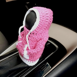 May include: Pink crocheted car gear shift cover with a white trim and a hood. The cover is shaped like a hooded sweatshirt.