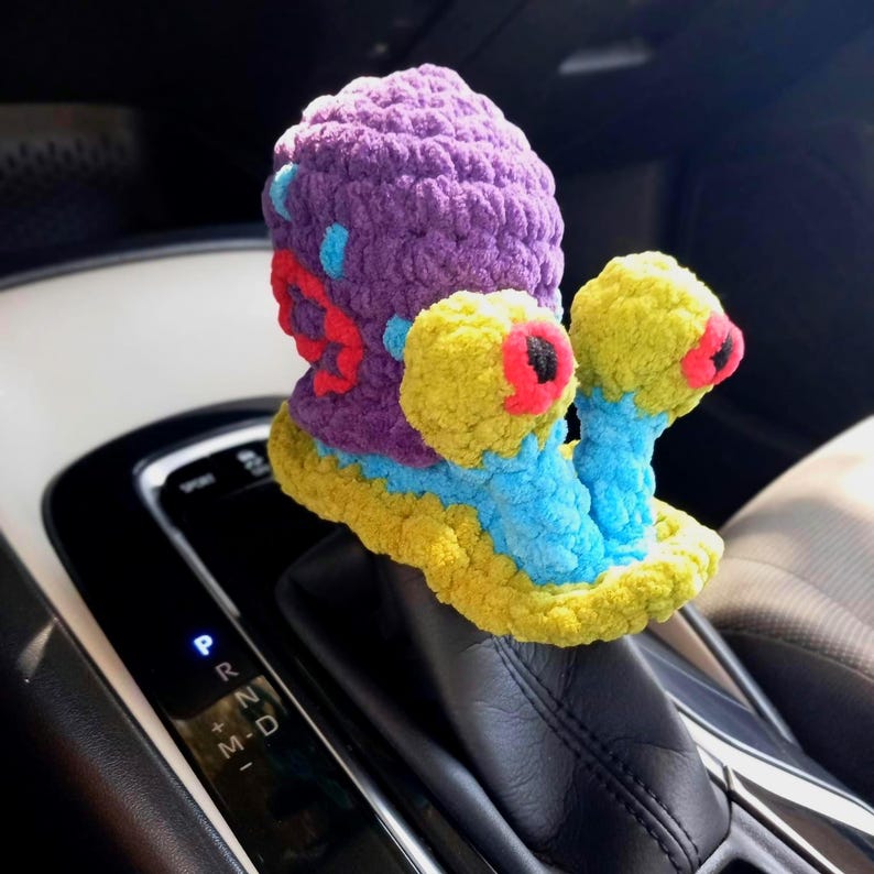 Pu&ograve; includere: Un accessorio per auto a forma di lumaca fatto a mano all'uncinetto. La lumaca presenta un corpo viola, dettagli gialli e blu e grandi occhi gialli con dettagli rossi e neri. La lumaca &egrave; posizionata sul cambio di un'auto.