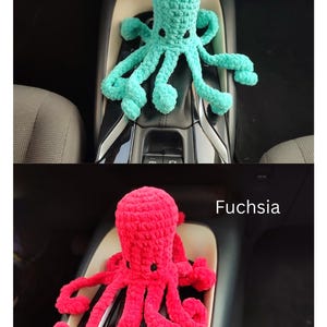 May include: Two crocheted octopus toys, one turquoise and one fuchsia, are displayed. The turquoise octopus is positioned above the fuchsia one. The text "Turquoise" and "Fuchsia" are above each octopus. The toys are placed in a car's console.