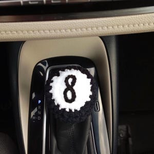 May include: A black and white crocheted shift knob cover featuring a number 8 design. The cover is circular with a white center and a black border. The number 8 is in black. The cover is placed over a car's gear shift.