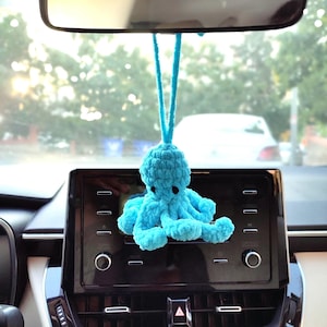 May include: A turquoise, crocheted octopus car charm suspended from a rearview mirror. The octopus features a round head, black eyes, and eight curled tentacles. The charm is hung by a matching turquoise cord, adding a playful element to the car's interior.
