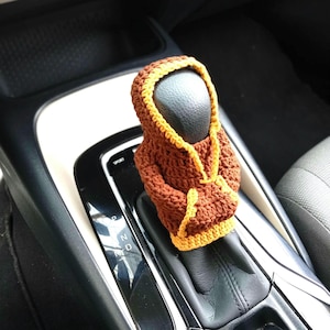 May include: A brown crocheted hoodie cover for a car gear shift knob, featuring a bright yellow trim along the hood opening, the cuffs, and the bottom hem. The garment is designed to fit snugly over the top of the black gear shifter handle.