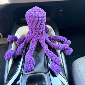 May include: A purple crocheted octopus plush toy with a textured, yarn-based surface and small black eyes. The octopus features a rounded head and eight long, curled tentacles that extend outward, resting on a dark surface with visible buttons.