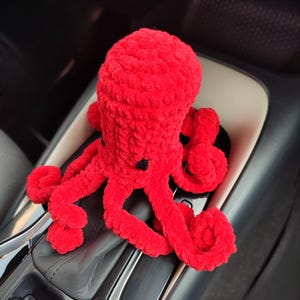 May include: A bright red, plush octopus toy with eight curled tentacles and small black eyes. The handmade stuffed animal is sitting on a car's console, showcasing its vibrant color and soft texture. A cute and cuddly plushie.