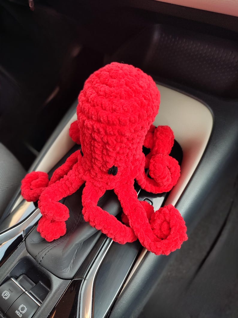 May include: A bright red, plush octopus toy with eight curled tentacles and small black eyes. The handmade stuffed animal is sitting on a car's console, showcasing its vibrant color and soft texture. A cute and cuddly plushie.