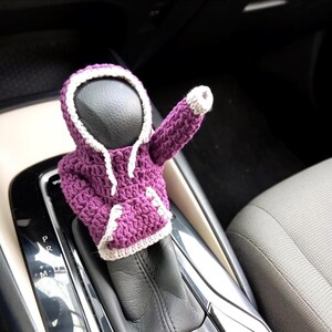 May include: Purple crocheted hoodie gear shift cover with white trim. The cover is on a car's gear shift lever.