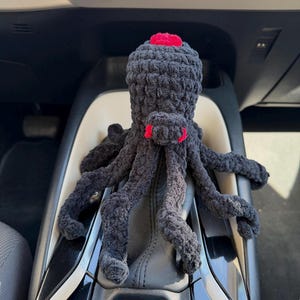 May include: A black crocheted octopus plush toy with a textured yarn surface, featuring a small red circular accent on the top of its head and two small red accents on the front of its body, positioned with eight dangling tentacles.