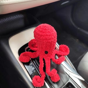 May include: A bright red, crocheted octopus toy with eight long, curled tentacles. The plush toy has a black button eye and is positioned on a car's gear shift. The background shows the car's interior, including the gear shift console and dashboard.