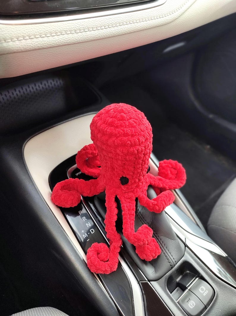 May include: A bright red, crocheted octopus toy with eight long, curled tentacles. The plush toy has a black button eye and is positioned on a car's gear shift. The background shows the car's interior, including the gear shift console and dashboard.