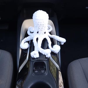 May include: A white crocheted octopus toy with eight tentacles. The plush toy is sitting on a black and silver car console. The octopus has a round head and is made of a soft, textured yarn.
