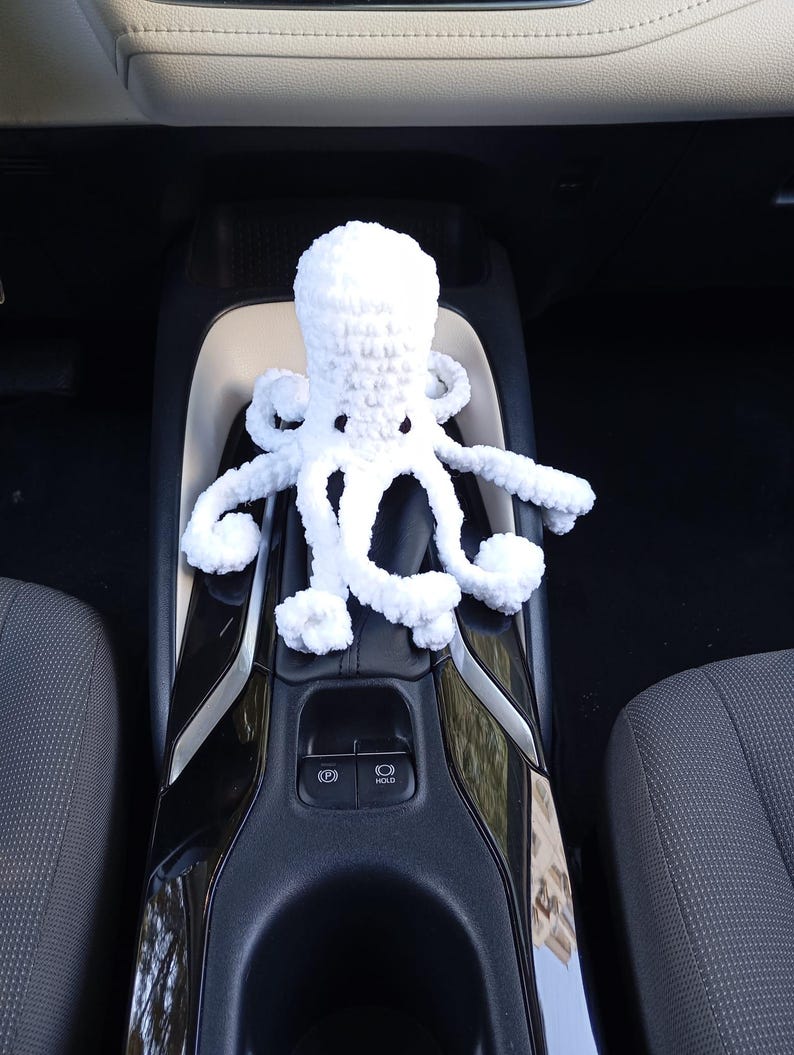 May include: A white crocheted octopus toy with eight tentacles. The plush toy is sitting on a black and silver car console. The octopus has a round head and is made of a soft, textured yarn.