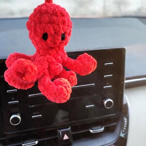 May include: A bright red, plush octopus car charm. The handmade octopus has a round head, eight curled tentacles, and small black eyes. The charm is hanging from a car dashboard.