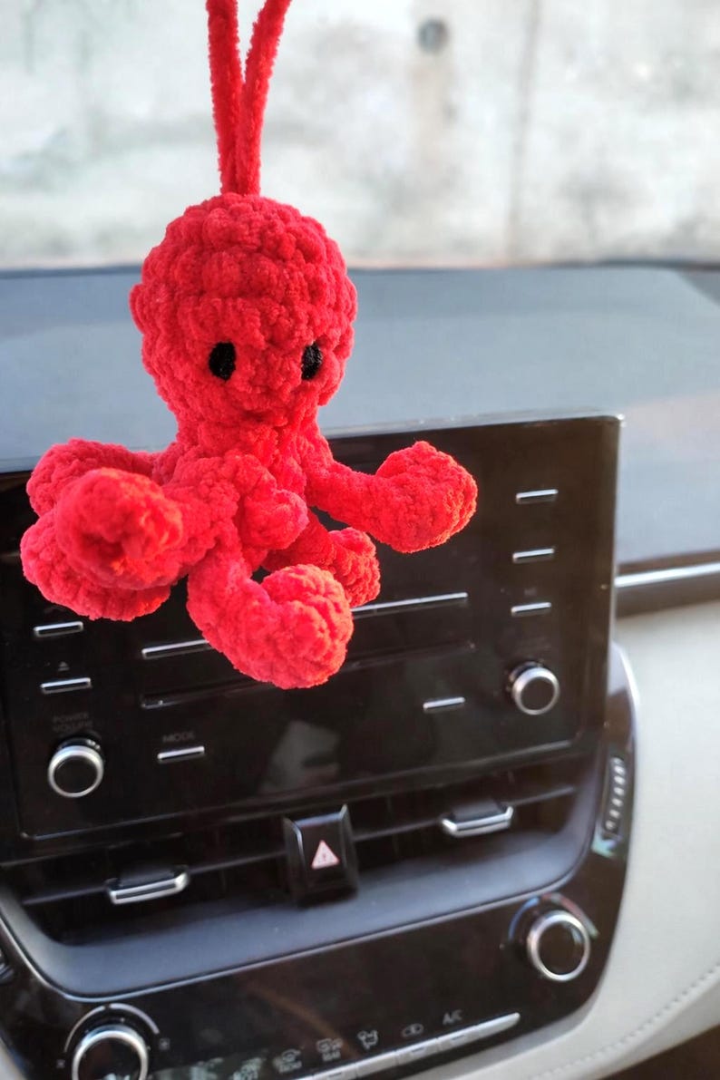 May include: A bright red, plush octopus car charm. The handmade octopus has a round head, eight curled tentacles, and small black eyes. The charm is hanging from a car dashboard.