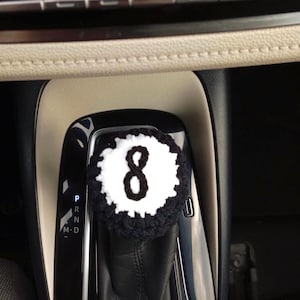Peut inclure: Une housse de levier de vitesse crochetée, ronde, conçue pour ressembler à une boule de billard numéro huit noire et blanche. La housse présente un centre circulaire blanc avec un chiffre huit noir cousu au milieu, bordé d'une épaisse bordure noire.