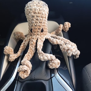 May include: A crocheted octopus in shades of beige and brown, with small black eyes. The plush toy has eight textured tentacles and is sitting on a car's console. The octopus is handmade and has a soft, cuddly appearance.