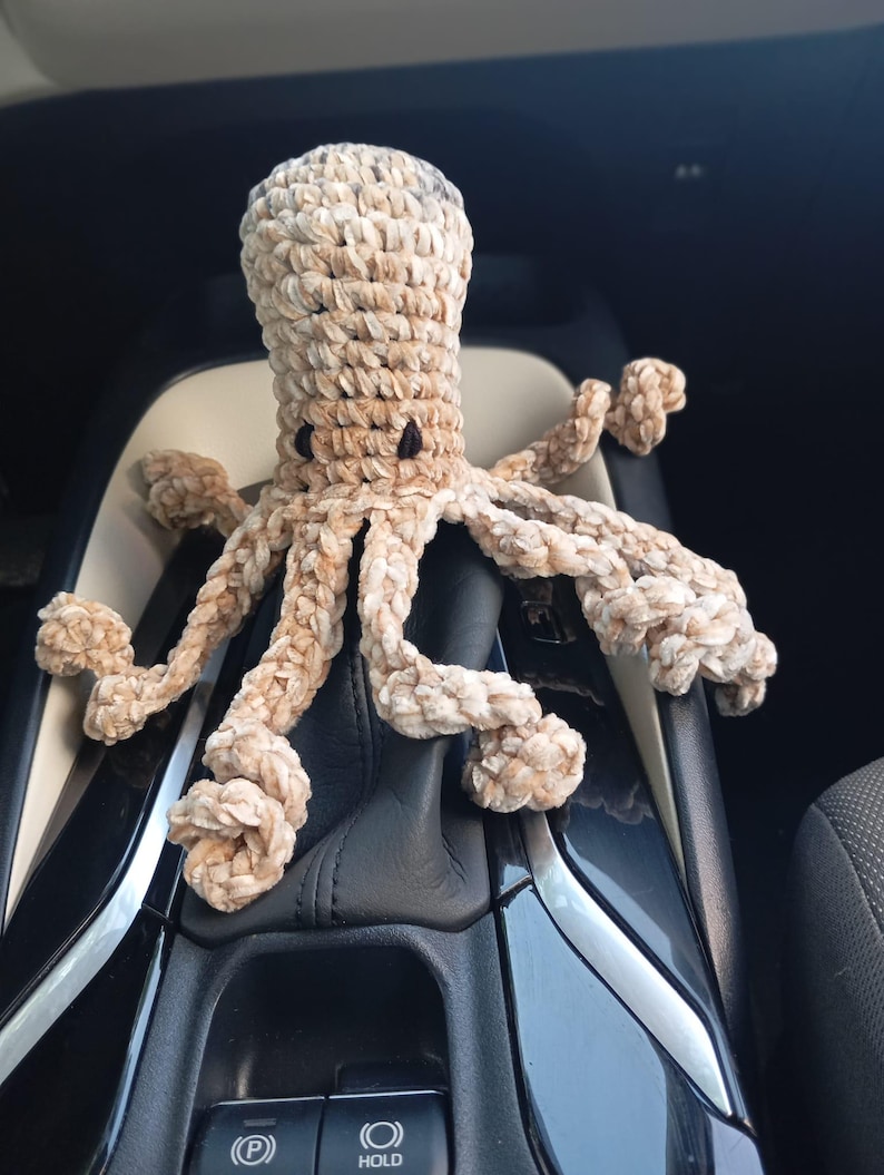 May include: A crocheted octopus in shades of beige and brown, with small black eyes. The plush toy has eight textured tentacles and is sitting on a car's console. The octopus is handmade and has a soft, cuddly appearance.